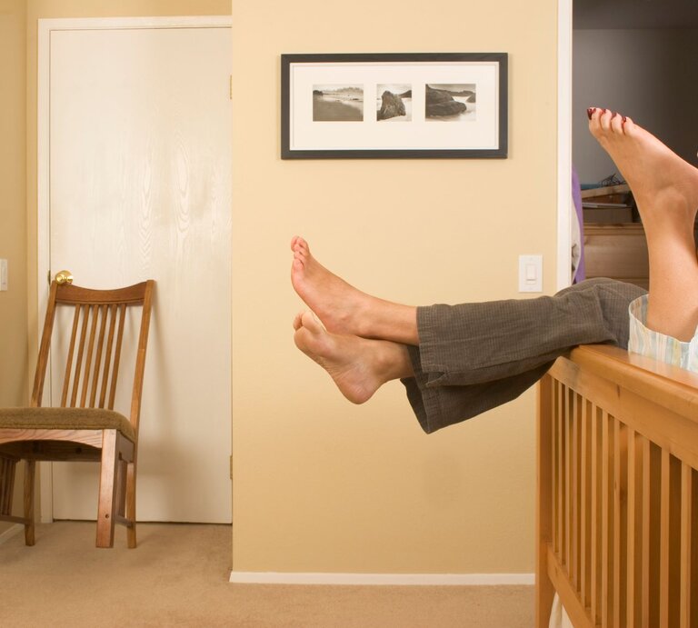 Zwei Paar nackte Beine, ein Mann in grauer Hose und eine Frau in Pyjama, ragen entspannt über ein Holzgeländer in einem hellen Schlafzimmer. | © Getty Images / Swell Media