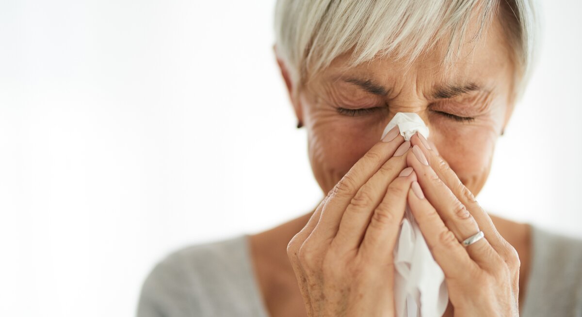 Eine Frau mit kurzen grauen Haaren benutzt ein Taschentuch, um ihre Nase zu putzen. Die Augen hat sie dabei geschlossen, was auf eine Erkältung oder eine Allergie hinweisen könnte. Die Frau steht vor einem unscharfen, hellen Hintergrund.