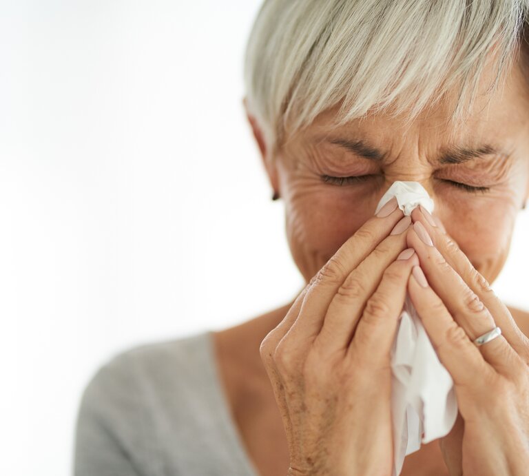 Eine Frau mit kurzen grauen Haaren benutzt ein Taschentuch, um ihre Nase zu putzen. Die Augen hat sie dabei geschlossen, was auf eine Erkältung oder eine Allergie hinweisen könnte. Die Frau steht vor einem unscharfen, hellen Hintergrund. | © iStock.com / Cecilie_Arcurs