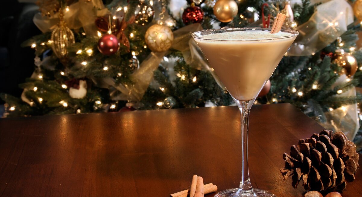 Eierlikör-Martini im Glas mit Zimtstange auf Holztisch mit Zimtstange, Haselnüssen und Tannenzapfen. Im Hintergrund Weihnachtsdekoration. Frontalansicht.