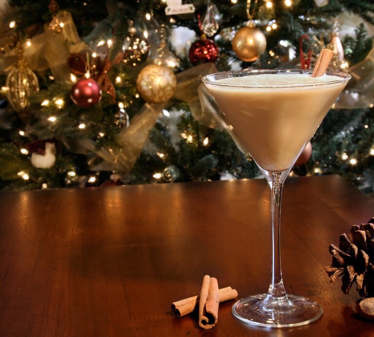 Eierlikör-Martini im Glas mit Zimtstange auf Holztisch mit Zimtstange, Haselnüssen und Tannenzapfen. Im Hintergrund Weihnachtsdekoration. Frontalansicht. | © Getty Images / MadCircles