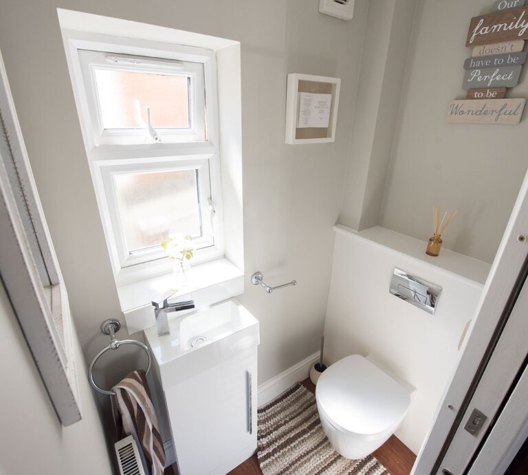 Ein kleines, helles Badezimmer mit weißer Einrichtung, ausgestattet mit einer Toilette, einem Waschbecken mit Spiegel darüber, einem Fenster, das Tageslicht hereinlässt, und dekorativen Elementen wie einem Bilderrahmen und einem Holzschild mit der Aufschrift "Our family doesn't have to be perfect to be wonderful". Ein gestreifter Teppich liegt auf dem Boden. | © Getty Images / John Keeble