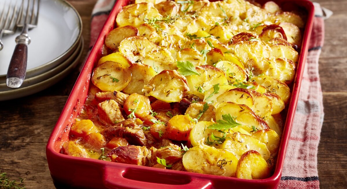 Putengulasch unter Kartoffelknusper-Haube in roter Auflaufform auf rotem Küchentuch, daneben Geschirr auf Holzuntergrund. 