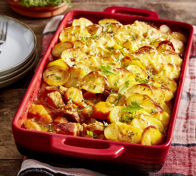 Putengulasch unter Kartoffelknusper-Haube in roter Auflaufform auf rotem Küchentuch, daneben Geschirr auf Holzuntergrund.  | © EAT CLUB / Isabell Triemer 