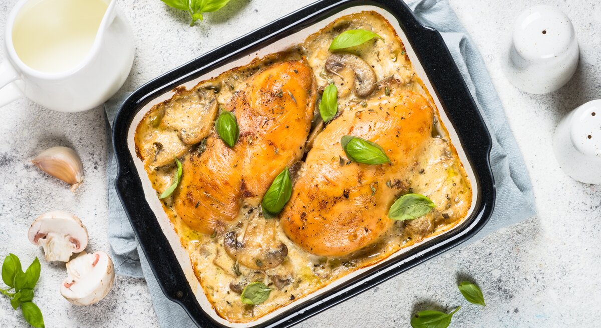 Draufsicht: heller Untergrund, eine Auflaufform mit Hühnerbrust mit Pilzen in Sahnesoße aus dem Ofen, sowie Pilze, Knoblauch Salz und Pfefferstreuer und ein Kännchen Milch.