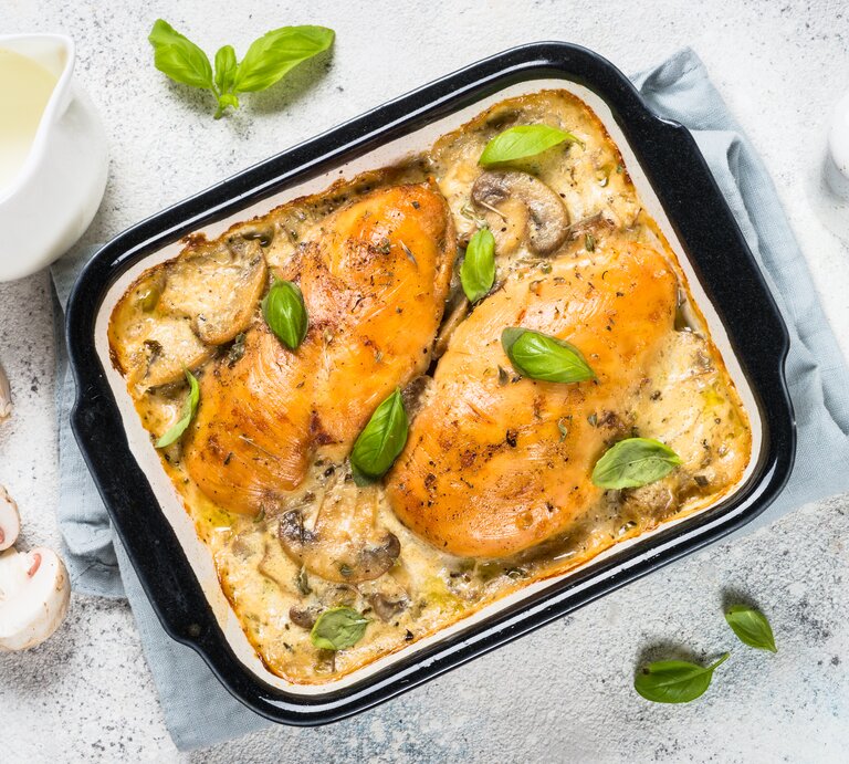 Draufsicht: heller Untergrund, eine Auflaufform mit Hühnerbrust mit Pilzen in Sahnesoße aus dem Ofen, sowie Pilze, Knoblauch Salz und Pfefferstreuer und ein Kännchen Milch. | © Shutterstock / nadianb