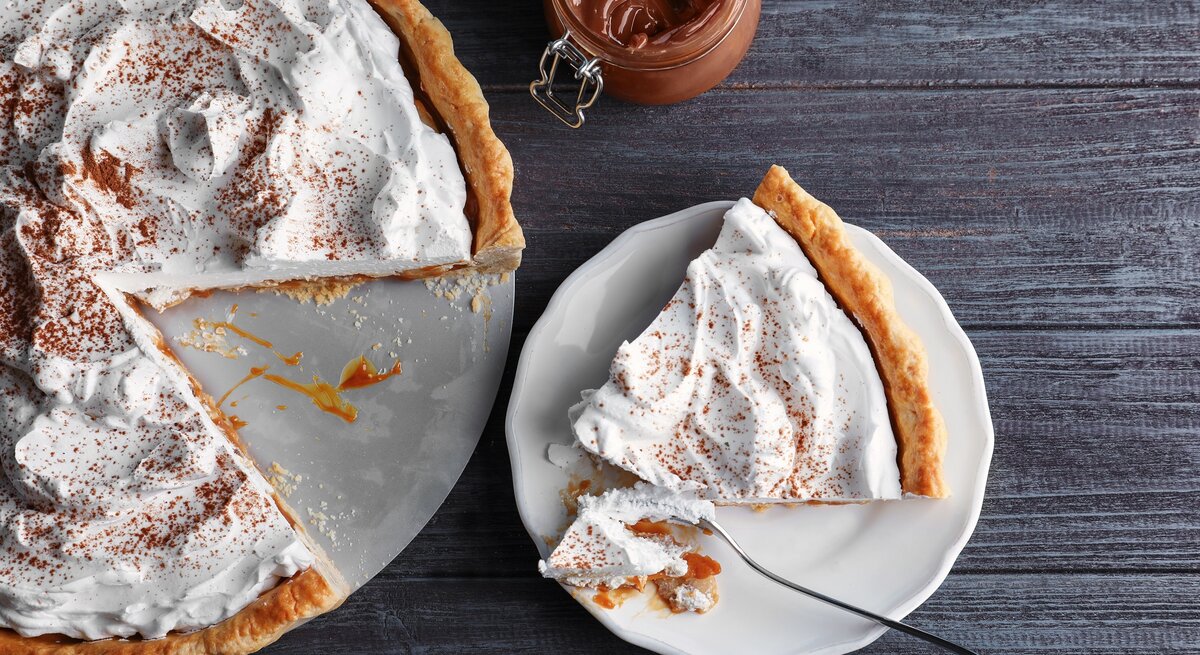 Ein angeschnittener Karamell-Kuchen mit Kokoscreme in der Draufsicht, daneben ein Weckglas mit Karamellsoße und ein Teller mit einem Stück des Kuchens.