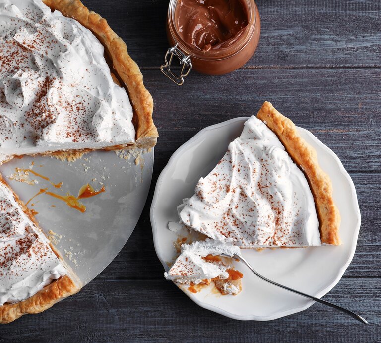 Ein angeschnittener Karamell-Kuchen mit Kokoscreme in der Draufsicht, daneben ein Weckglas mit Karamellsoße und ein Teller mit einem Stück des Kuchens. | © Adobe Stock/ Africa Studio