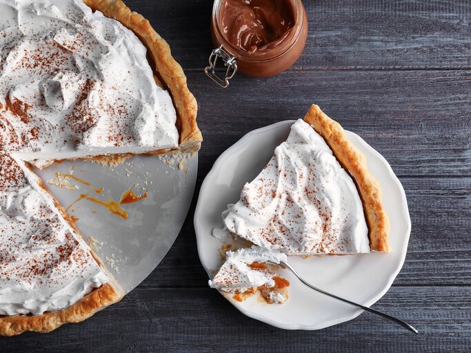 Ein angeschnittener Karamell-Kuchen mit Kokoscreme in der Draufsicht, daneben ein Weckglas mit Karamellsoße und ein Teller mit einem Stück des Kuchens. | © Adobe Stock/ Africa Studio