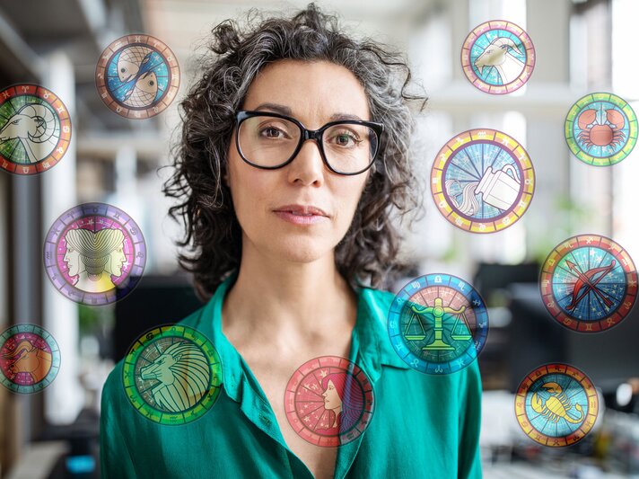 Eine Frau mit lockigen grauen Haaren und Brille steht in einem Büro. Die astrologischen Symbole der 12 Sternzeichen umgeben sie. | © iStock [M]