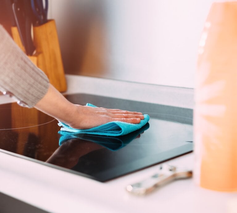 Person, die eine Küchenarbeitsplatte mit einem blauen Mikrofasertuch reinigt. Der Fokus liegt auf der Hand der Person, die das Tuch über die glänzende, schwarze Oberfläche eines Kochfeldes führt. | © GettyImages / Cris Cantón