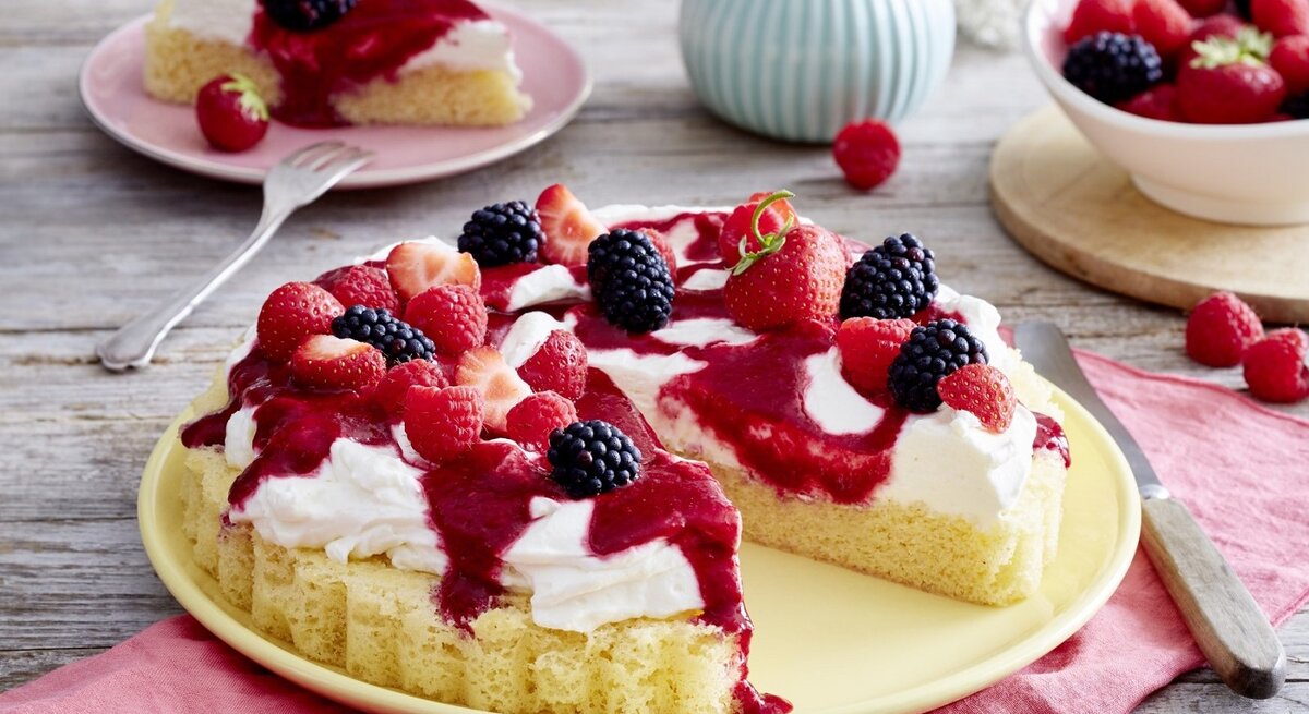 Auf einem Teller steht ein angeschnittener Schmand-Obstkuchen. Er ist mit Himbeersauce und Beeren überzogen. 