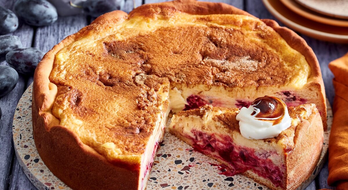Angeschnittener Pfälzer-Zwetschgen-Cheesecake mit Sahne auf einer runden Unterlage, Holzuntergrund und im Hintergrund Zwetschgen und Teller. 