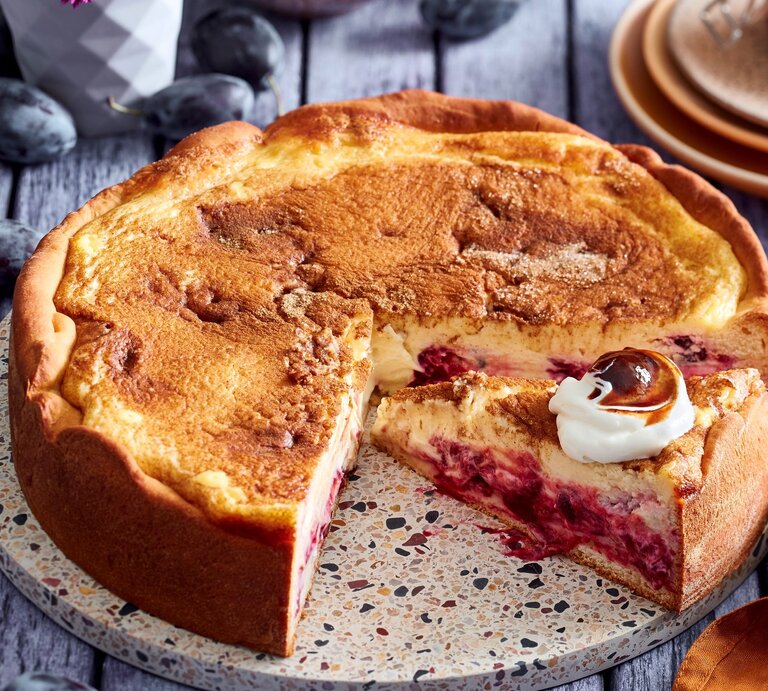 Angeschnittener Pfälzer-Zwetschgen-Cheesecake mit Sahne auf einer runden Unterlage, Holzuntergrund und im Hintergrund Zwetschgen und Teller.  | © EAT CLUB / Isabell Triemer