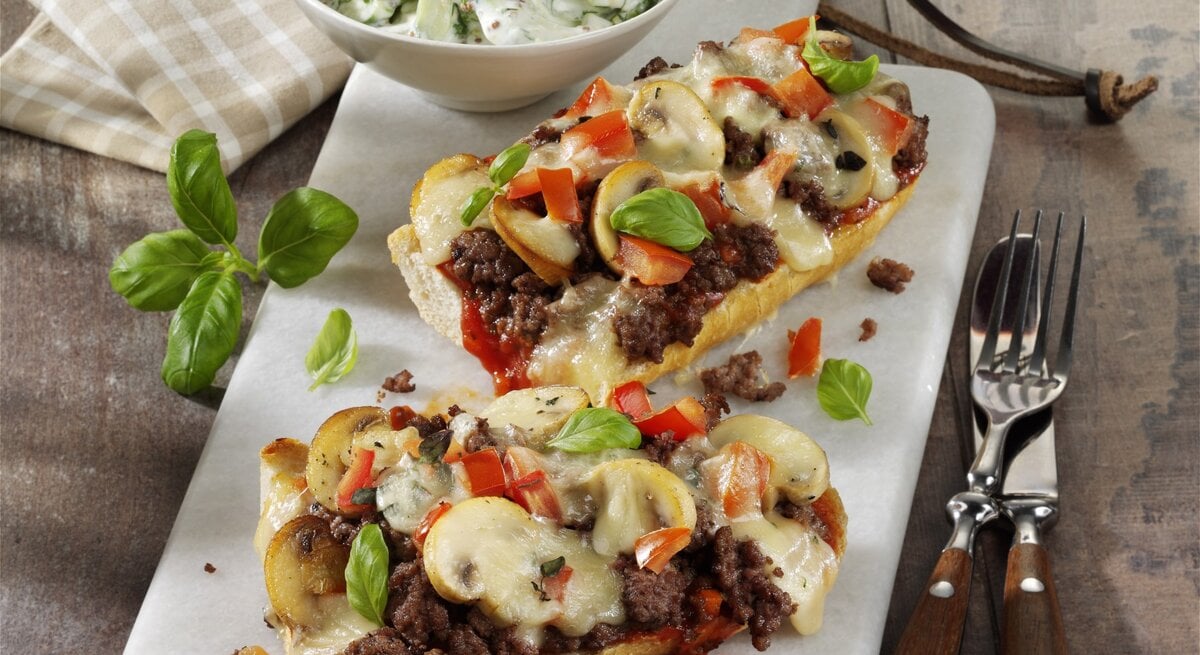 Französische Brotpizza mit Hackfleisch und Pilzen, garniert mit frischer Paprika und Basilikum und serviert mit einem frischen selbst gemachten Tzaziki auf einer weißen Steinplatte auf hölzernem Untergrund. 