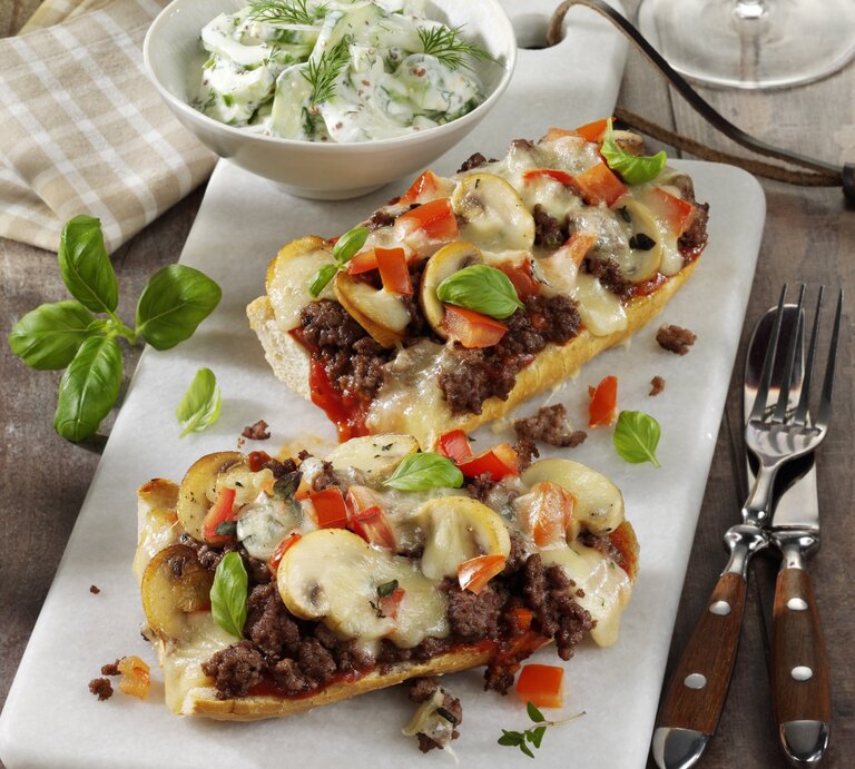 Französische Brotpizza mit Hackfleisch und Pilzen, garniert mit frischer Paprika und Basilikum und serviert mit einem frischen selbst gemachten Tzaziki auf einer weißen Steinplatte auf hölzernem Untergrund.  | © StockFood / StockFood Studios / Photoart