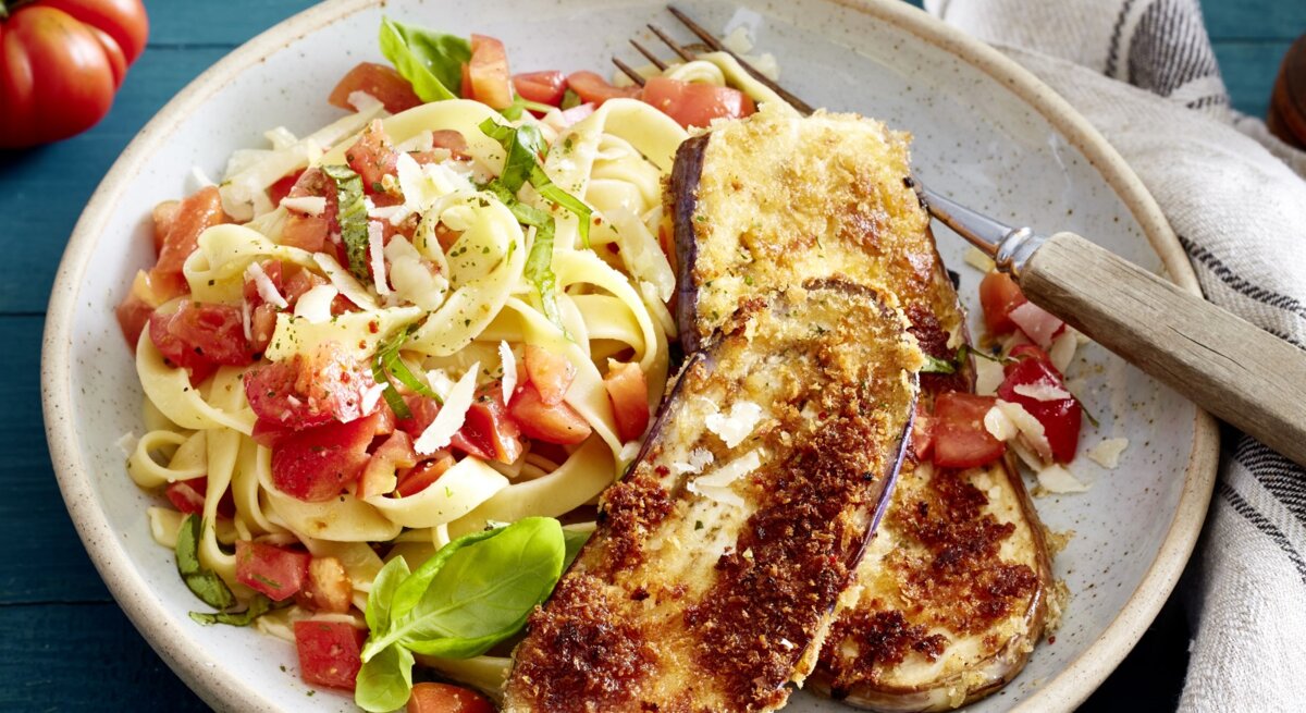 Auberginen-Schnitzel mit Bruschetta-Nudeln und Gabel auf einem hellen Teller. Drum herum Basilikum, Tomaten und ein Küchentuch, alles auf blauem Untergrund. 