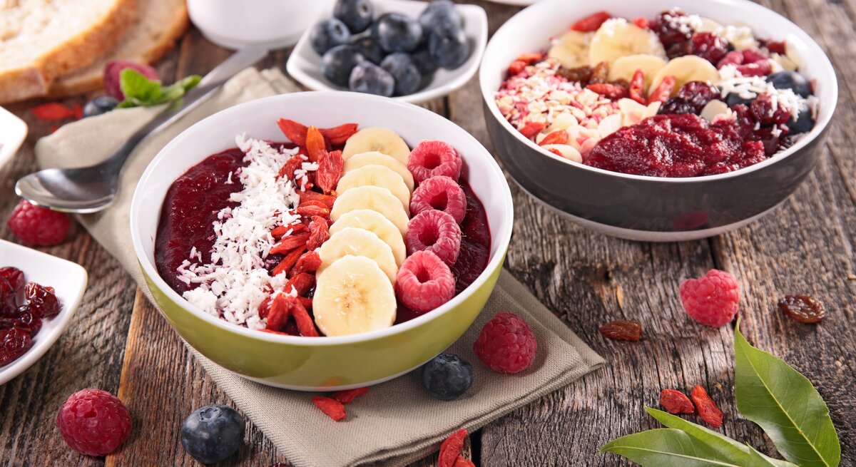 Ein gesundes Frühstück ist auf einem rustikalen Holztisch angerichtet: Zwei Schüsseln mit Acai-Beeren-Smoothie, garniert mit Bananenscheiben, Himbeeren, Kokosraspeln und Goji-Beeren. Daneben sind Schalen mit frischen Blaubeeren und Himbeeren, ein Stück Brot, eine Tasse Kaffee und einige Blätter zu sehen. Der Tisch präsentiert eine reichhaltige Auswahl an farbenfrohen und nahrhaften Lebensmitteln, die eine gesunde Mahlzeit darstellen.
