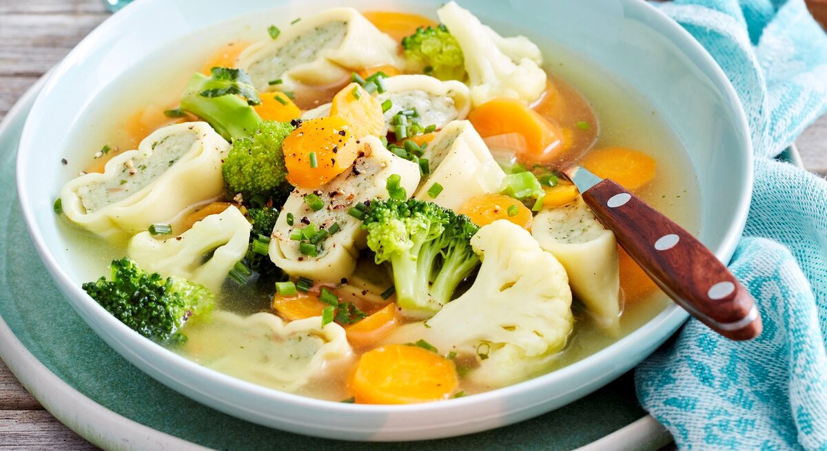 Weiße Schüssel mit Gemüsetopf mit Maultaschen und Löffel vor Holzhintergrund. 