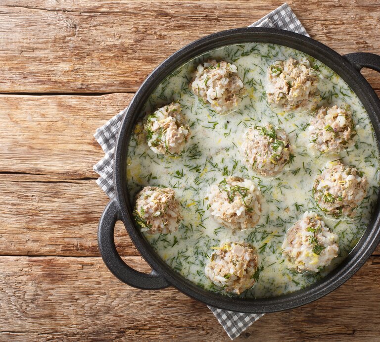Saftige Reis-Hackbällchen serviert in cremiger Dill-Sahne-Soße in einer schwarzen gusseisernen Form. Das Ganze steht auf einem mit einem karierten Tischtuch bedecktem Holztisch. | © Adobe-Stock/FomaA