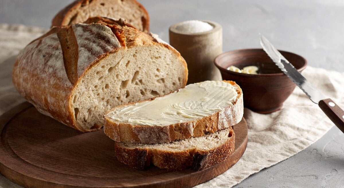 Ein angeschnittener Laib "Friss dich dumm"-Brot auf einem Brett, zwei Scheiben liegen davor, eine Scheibe mit Butter bestrichen, daneben eine Schale Butter mit einem Messer und eine Schale Salz.
