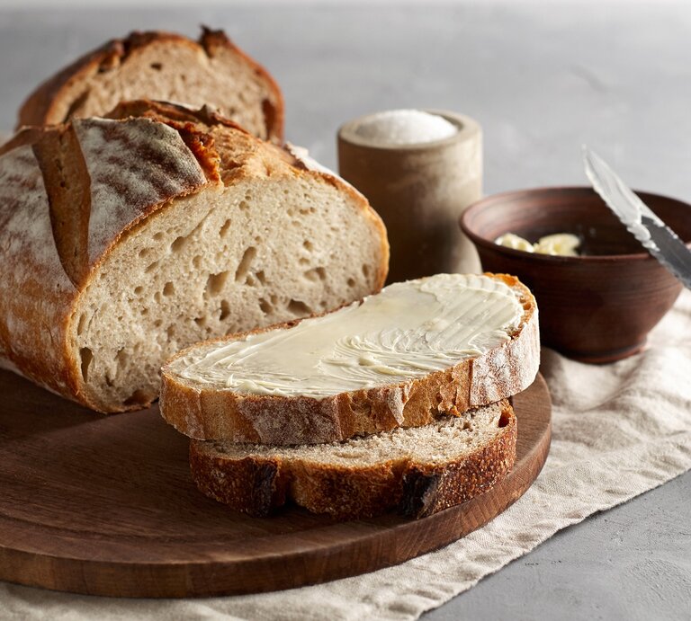 Ein angeschnittener Laib "Friss dich dumm"-Brot auf einem Brett, zwei Scheiben liegen davor, eine Scheibe mit Butter bestrichen, daneben eine Schale Butter mit einem Messer und eine Schale Salz. | © Adobe Stock/ Dariia Belkina