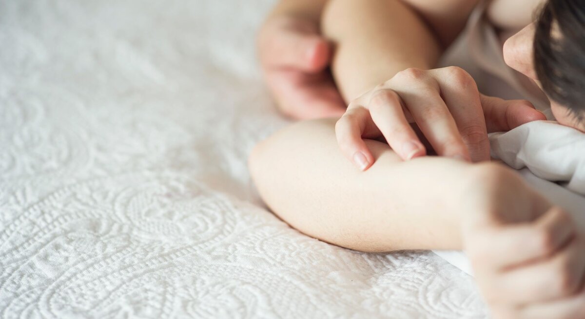 Paar liegt zusammen im Bett. Zu sehen ist seine Hand, die auf ihrer Hand liegt.