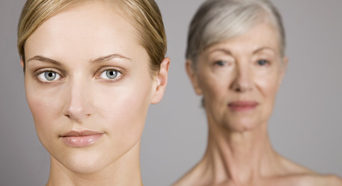 Zwei Frauen in einem Bild, die jüngere im Vordergrund mit Blick in die Kamera und die ältere im unscharfen Hintergrund, symbolisieren verschiedene Generationen oder den Alterungsprozess.