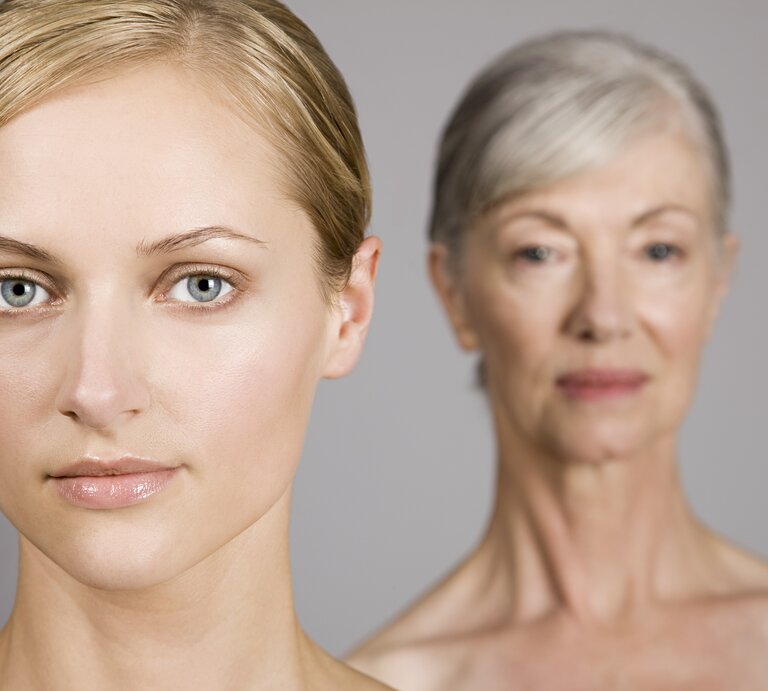 Zwei Frauen in einem Bild, die jüngere im Vordergrund mit Blick in die Kamera und die ältere im unscharfen Hintergrund, symbolisieren verschiedene Generationen oder den Alterungsprozess. | © Getty Images/Image Source