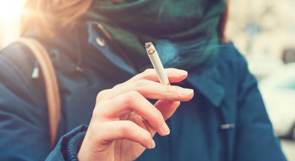 Eine junge Frau hält eine brennende Zigarette zwischen Zeige- und Mittelfinger. An den Stellen, an denen die Zigarette die Haut berührt, werden sich mit der Zeit gelbe Flecken bilden. Wie wird man sie los?