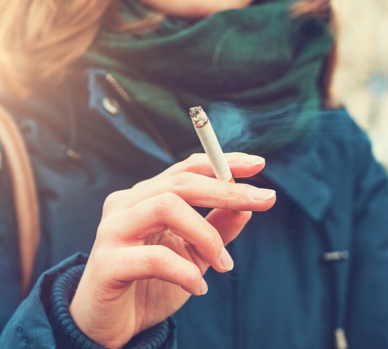 Eine junge Frau hält eine brennende Zigarette zwischen Zeige- und Mittelfinger. An den Stellen, an denen die Zigarette die Haut berührt, werden sich mit der Zeit gelbe Flecken bilden. Wie wird man sie los? | © iStock.com/Terroa