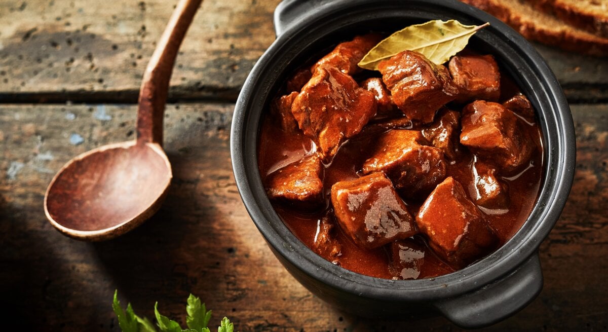 Gulasch in einem kleinen schwarzen Tontopf auf einem rustikalen Holztisch mit einem Holzkochlöffel und Petersilie.