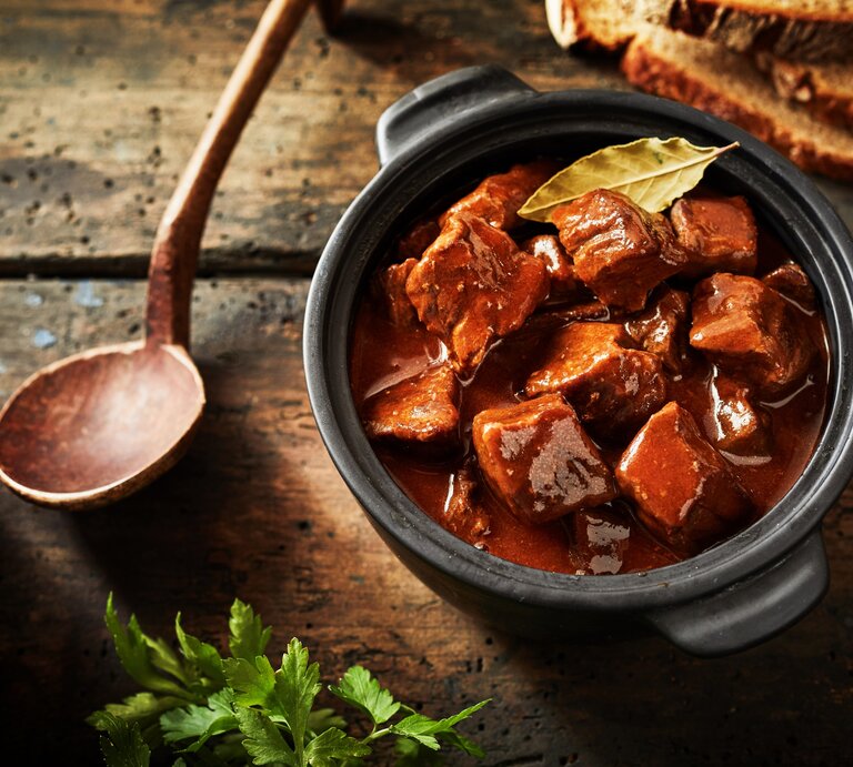 Gulasch in einem kleinen schwarzen Tontopf auf einem rustikalen Holztisch mit einem Holzkochlöffel und Petersilie. | © Shuttertsock/ stockcreations