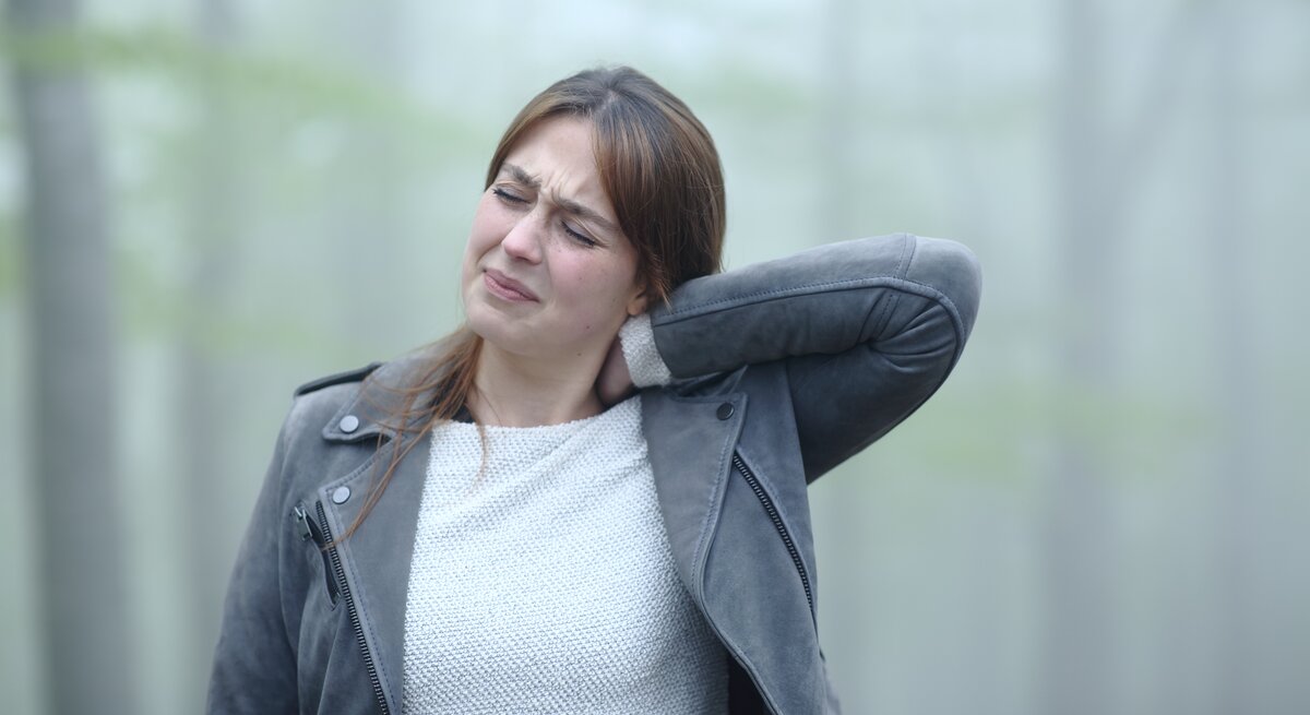 Eine junge Frau mit schmerzverzerrtem Gesicht hält sich den Nacken, als ob sie starke Schmerzen hätte. Sie trägt eine graue Lederjacke und einen hellen Strickpullover. Der Hintergrund ist verschwommen, mit sanften grünen Farbtönen, was auf eine Außenaufnahme hindeutet.