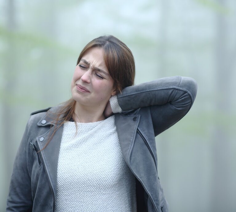 Eine junge Frau mit schmerzverzerrtem Gesicht hält sich den Nacken, als ob sie starke Schmerzen hätte. Sie trägt eine graue Lederjacke und einen hellen Strickpullover. Der Hintergrund ist verschwommen, mit sanften grünen Farbtönen, was auf eine Außenaufnahme hindeutet. | © Getty Images/AntonioGuillem