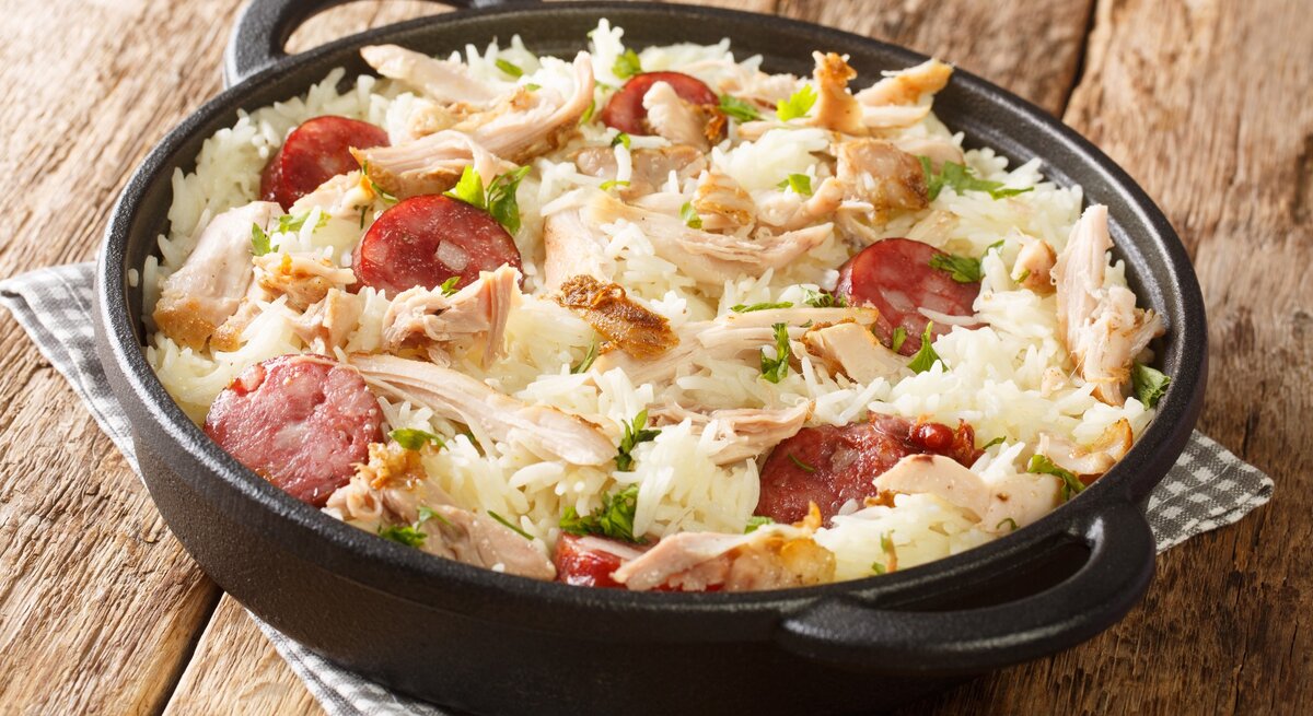 One Pot Reis mit Huhn und gebratener Wurst in Gusspfanne auf Stoffserviette auf Holztisch. Aufnahme aus der Draufsicht.