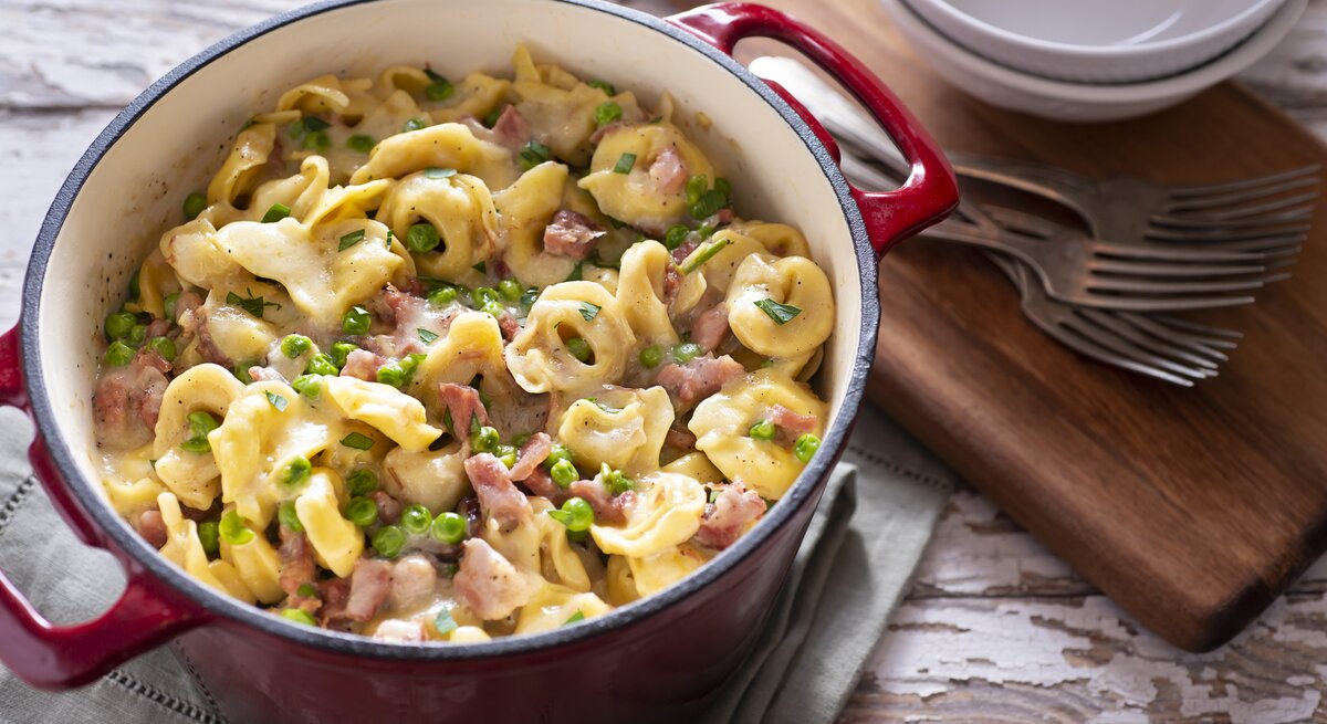 Tortellini mit Schinken und Erbsen in Sahnesoße, angerichtet in einem Topf. Daneben finden sich Holzbrett, Schalen und Gabeln. 