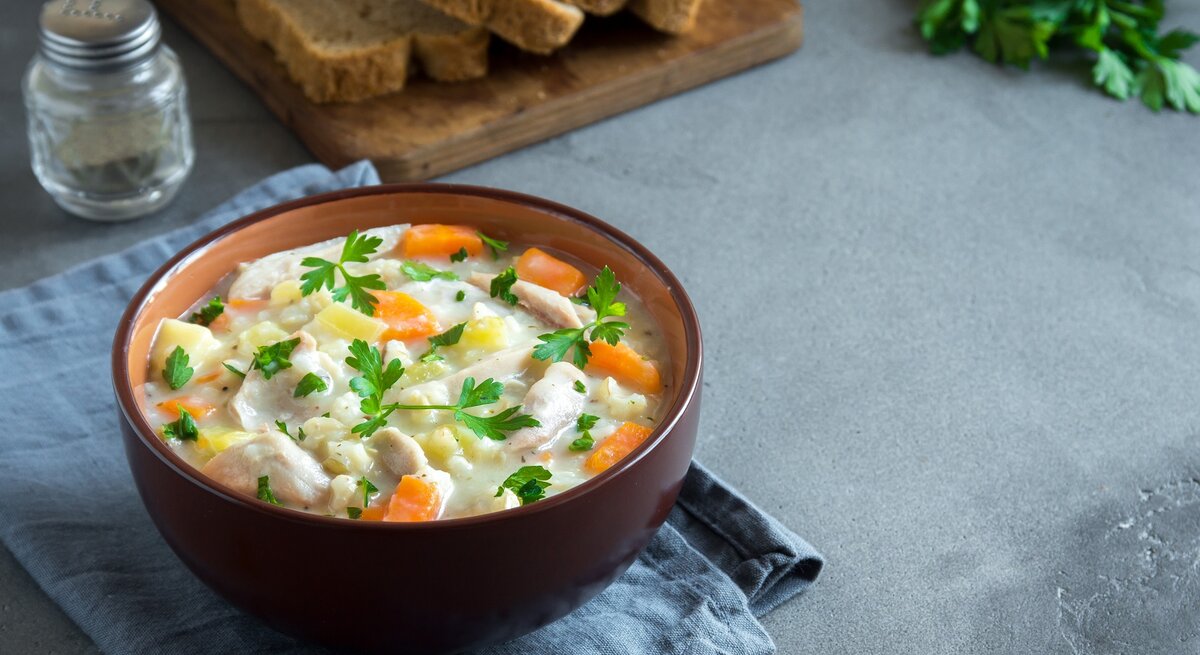 Eine braune Schale mit der Hühnersuppe auf grauem Untergrund. Im Hintergrund Petersilie, Brotscheiben und Pfeffer.
