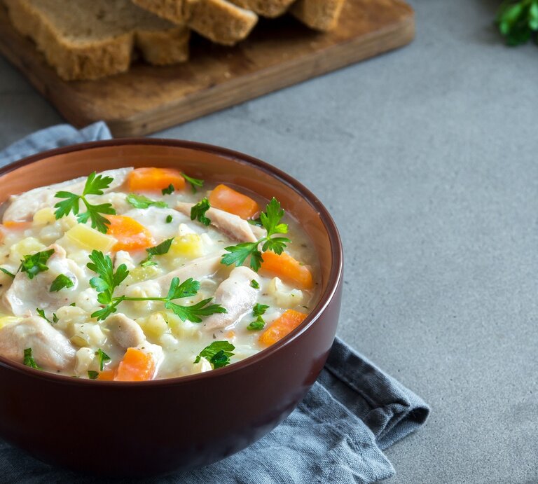 Eine braune Schale mit der Hühnersuppe auf grauem Untergrund. Im Hintergrund Petersilie, Brotscheiben und Pfeffer. | © Shutterstock/ Oksana Mizina
