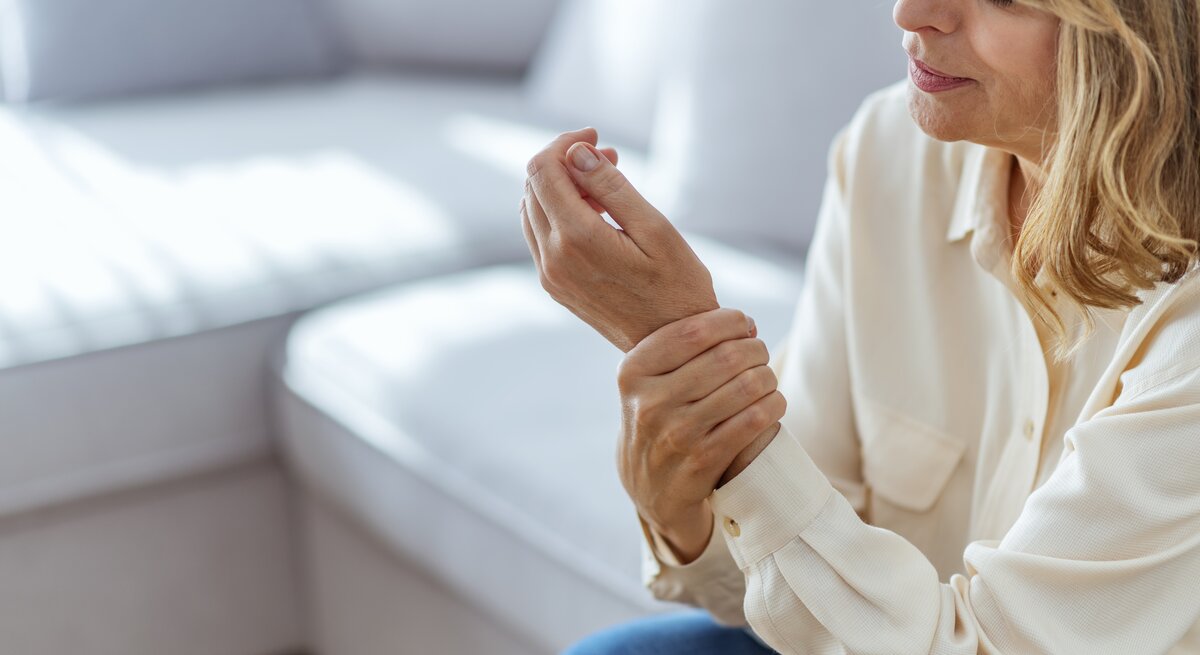 Eine Frau mittleren Alters in einem hellen, lässigen Hemd sitzt und betrachtet besorgt ihr schmerzendes Handgelenk. Sie hält mit ihrer anderen Hand das Handgelenk, was auf Unbehagen oder Schmerzen hindeutet. Ihr Gesichtsausdruck spiegelt Sorge und Unbehagen wider. Sie befindet sich in einem hellen Raum mit weicher Einrichtung im Hintergrund, was auf eine häusliche oder möglicherweise medizinische Umgebung hindeutet.