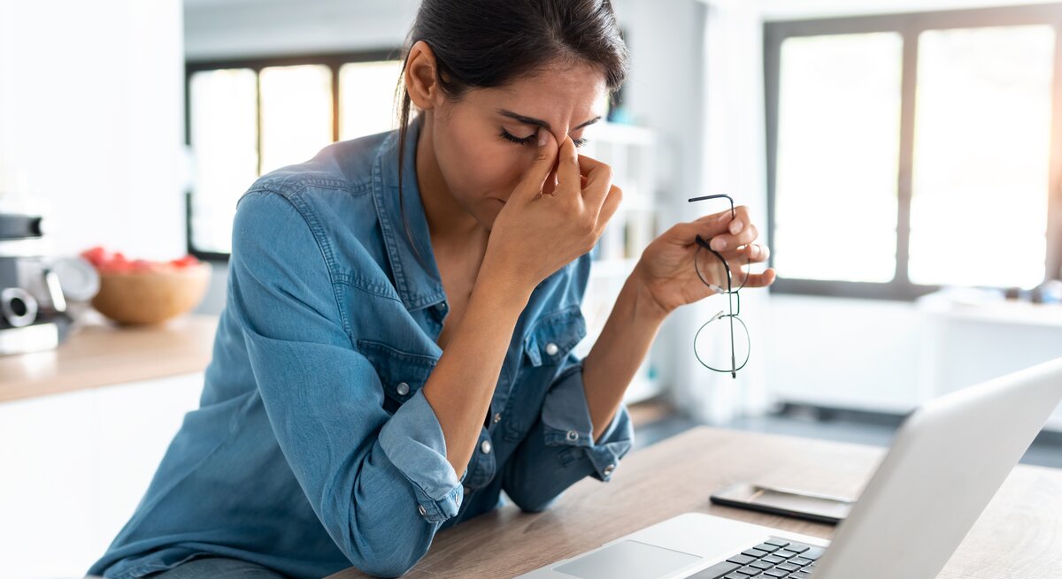Eine Frau in einem blauen Jeanshemd sitzt vor einem Laptop und hält ihre Brille in einer Hand, während sie mit der anderen Hand ihre Nasenwurzel massiert, ein Zeichen von Stress oder Ermüdung. Vor ihr auf dem Tisch liegen ein Notizblock und ein Smartphone. Der Raum ist hell und modern eingerichtet, mit großen Fenstern, die für natürliches Licht sorgen. Die Atmosphäre scheint ruhig, doch die Frau wirkt nachdenklich oder gestresst.