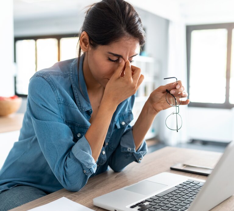 Eine Frau in einem blauen Jeanshemd sitzt vor einem Laptop und hält ihre Brille in einer Hand, während sie mit der anderen Hand ihre Nasenwurzel massiert, ein Zeichen von Stress oder Ermüdung. Vor ihr auf dem Tisch liegen ein Notizblock und ein Smartphone. Der Raum ist hell und modern eingerichtet, mit großen Fenstern, die für natürliches Licht sorgen. Die Atmosphäre scheint ruhig, doch die Frau wirkt nachdenklich oder gestresst. | © Getty Imges/nensuria