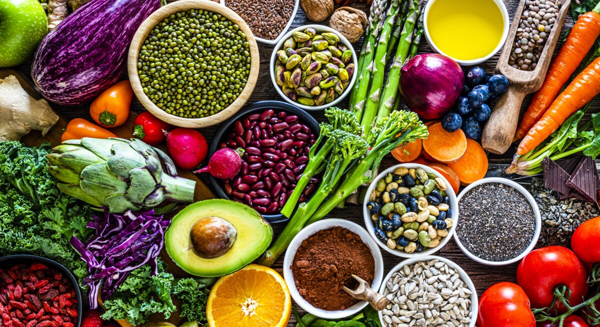 Ein reichhaltiges und farbenfrohes Arrangement von Superfoods und gesunden Lebensmitteln auf einem rustikalen Holztisch. Zu den sichtbaren Lebensmitteln gehören verschiedene frische Gemüsesorten wie Auberginen, Tomaten und grünes Blattgemüse, Obst wie Äpfel und Beeren, Nüsse, Samen, Hülsenfrüchte und Gewürze. Das Bild strahlt Frische und Vielfalt aus und betont eine nährstoffreiche Ernährung.