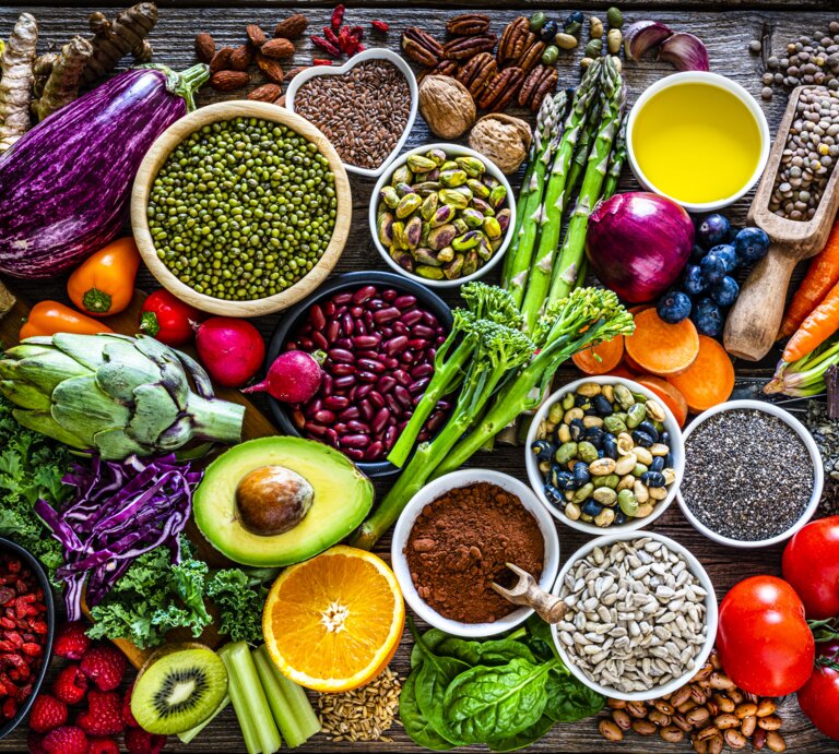 Ein reichhaltiges und farbenfrohes Arrangement von Superfoods und gesunden Lebensmitteln auf einem rustikalen Holztisch. Zu den sichtbaren Lebensmitteln gehören verschiedene frische Gemüsesorten wie Auberginen, Tomaten und grünes Blattgemüse, Obst wie Äpfel und Beeren, Nüsse, Samen, Hülsenfrüchte und Gewürze. Das Bild strahlt Frische und Vielfalt aus und betont eine nährstoffreiche Ernährung. | © Getty Images/fcafotodigital