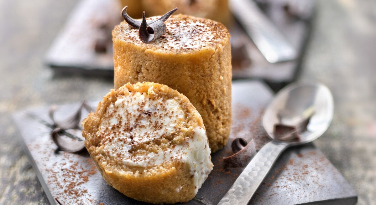 Tiramisu-Röllchen auf einem quadratischen schwarzen Brett. Daneben liegt ein Löffel. 