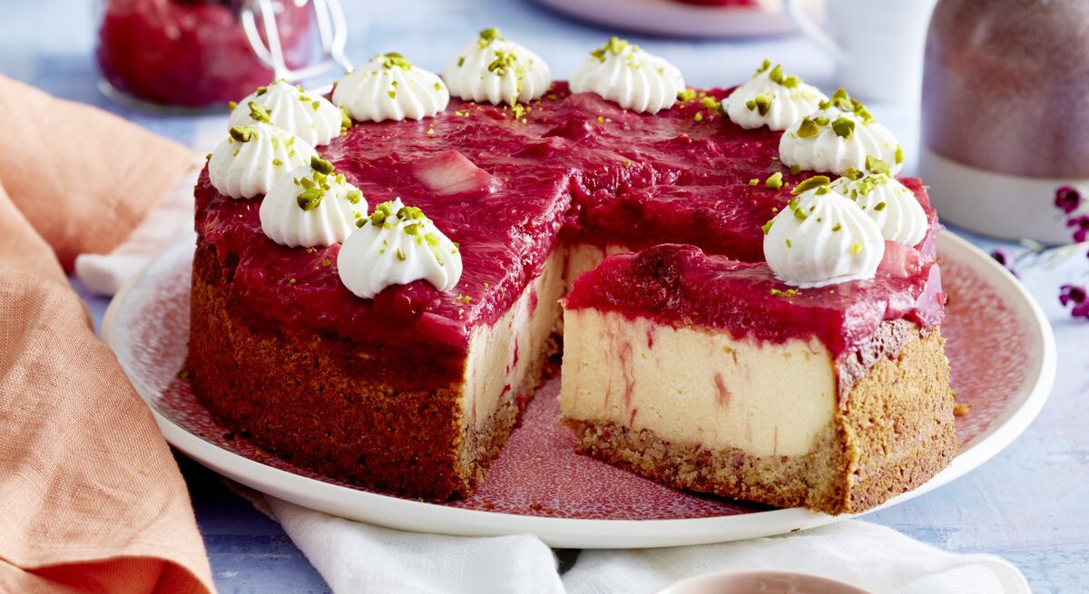 Auf einem Teller ist ein Rhabarber-Grieß-Cheesecake angerichtet. Er ist mit Sahnetüpfeln und Pistazien dekoriert.