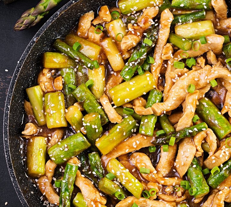 Eine schwarze Pfanne auf dunklem Untergrund mit grünem Spargel und Sesam direkt von oben fotografiert. | © Shutterstock/ Timolina