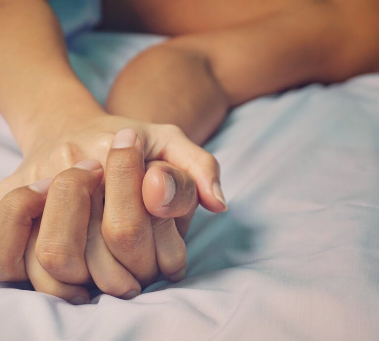 Zwei Hände, die sich fest auf einer blauen Bettdecke halten, wobei eine Person im Hintergrund undeutlich zu sehen ist, was Intimität und Nähe suggeriert. | © iStock / Napadon Srisawang