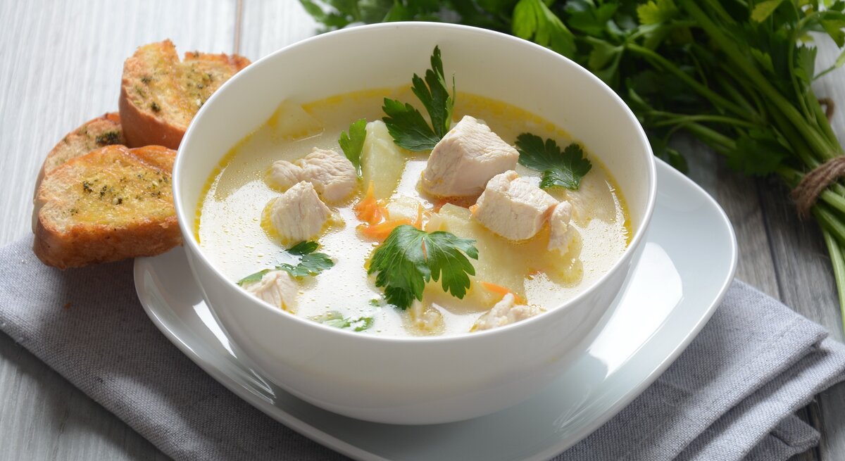 Eine weiße Suppenschale mit der Hühnerfrikassee-Suppe auf einem hellen Holztisch seitlich von schräg-oben fotografiert. Daneben noch ein Bund frische Petersilie auf der linken Seite.