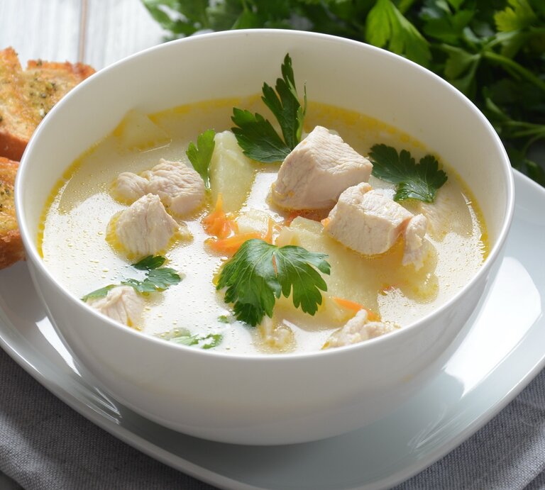 Eine weiße Suppenschale mit der Hühnerfrikassee-Suppe auf einem hellen Holztisch seitlich von schräg-oben fotografiert. Daneben noch ein Bund frische Petersilie auf der linken Seite. | © Shutterstock/ Natalia Hanin 